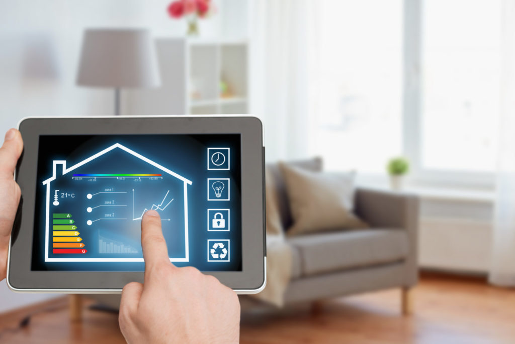 Haende einer Person halten ein Tablet in der Hand. Auf dem Tablet ist ein symbolisierter Querschnitt eines Hauses vor blauem Hintergrund. In dem Haus sind die verschiedenen Energieeffizienzklassen sowie eine Temperaturanzeige, ein Graph, ein Diagramm und eine Leiste mit verschiedenen Temperaturen von dunkelblau bis dunkelrot abgebildet. Die Person deutet mit dem Zeigefinger der rechten Hand auf den Graphen. Neben dem Haus sind vier weitere Symbole jeweils in einem quadratischen kleinen Kasten abgebildet. Eine Uhr, eine Gluehlampe, ein Schloss und das Symbol fuer Recycling. Im Hintergrund hinter dem Tablet ist ein Raum mit einem hellgrauen Sofa, einer weißen Stehlampe und einem Regal.