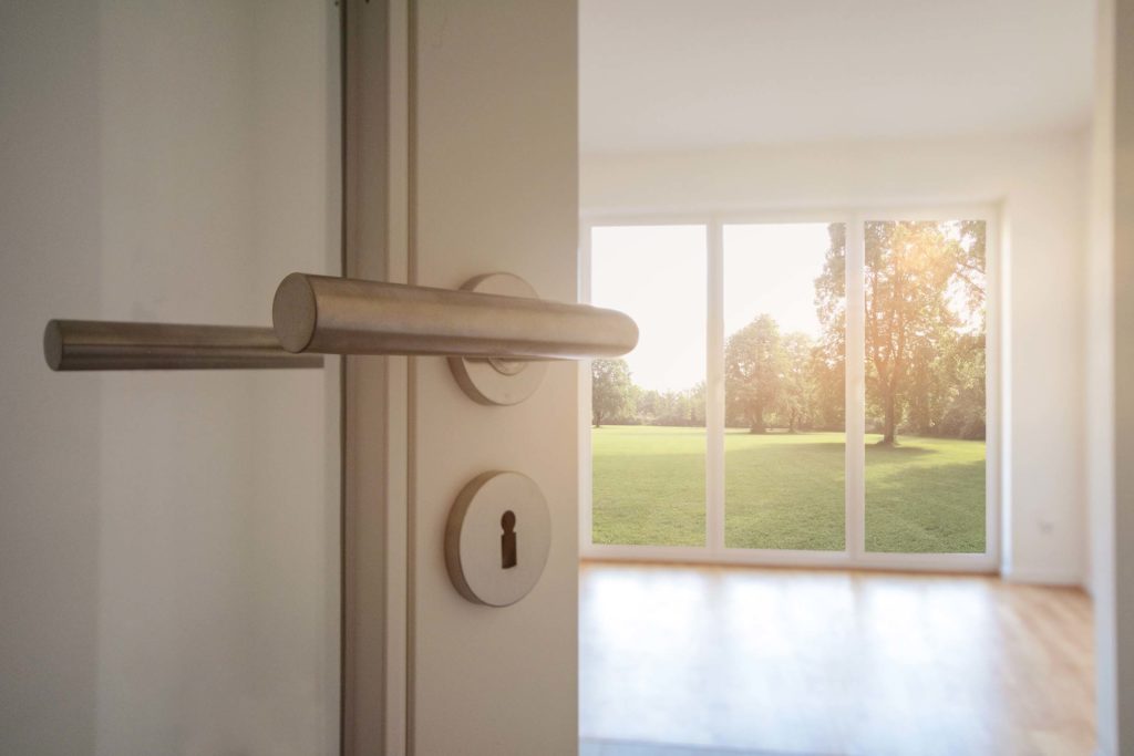 Blick durch eine halboffene, weiße Tuer auf Hoehe der Klinke in ein lichtdurchflutetes Zimmer mit bodentiefen Fenstern. Das Zimmer ist leer. Durch die Fenster ist eine große Rasenflaeche mit vielen gruenen Baeumen zu sehen.