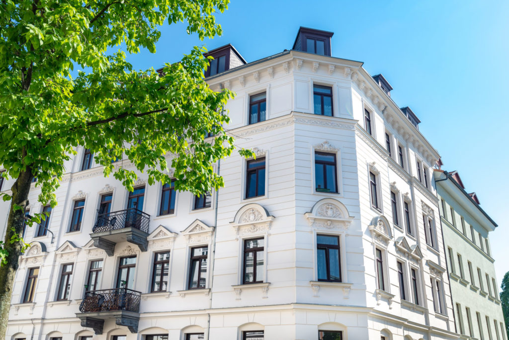 Leicht seitliche Frontansicht eines weißen Mehrfamilienhauses mit schwarzen Fensterrahmen und Stuckverzierung. Der Himmel ueber dem Haus ist hellblau und vor dem Haus ragt ein großer gruener Baum ins Bild.