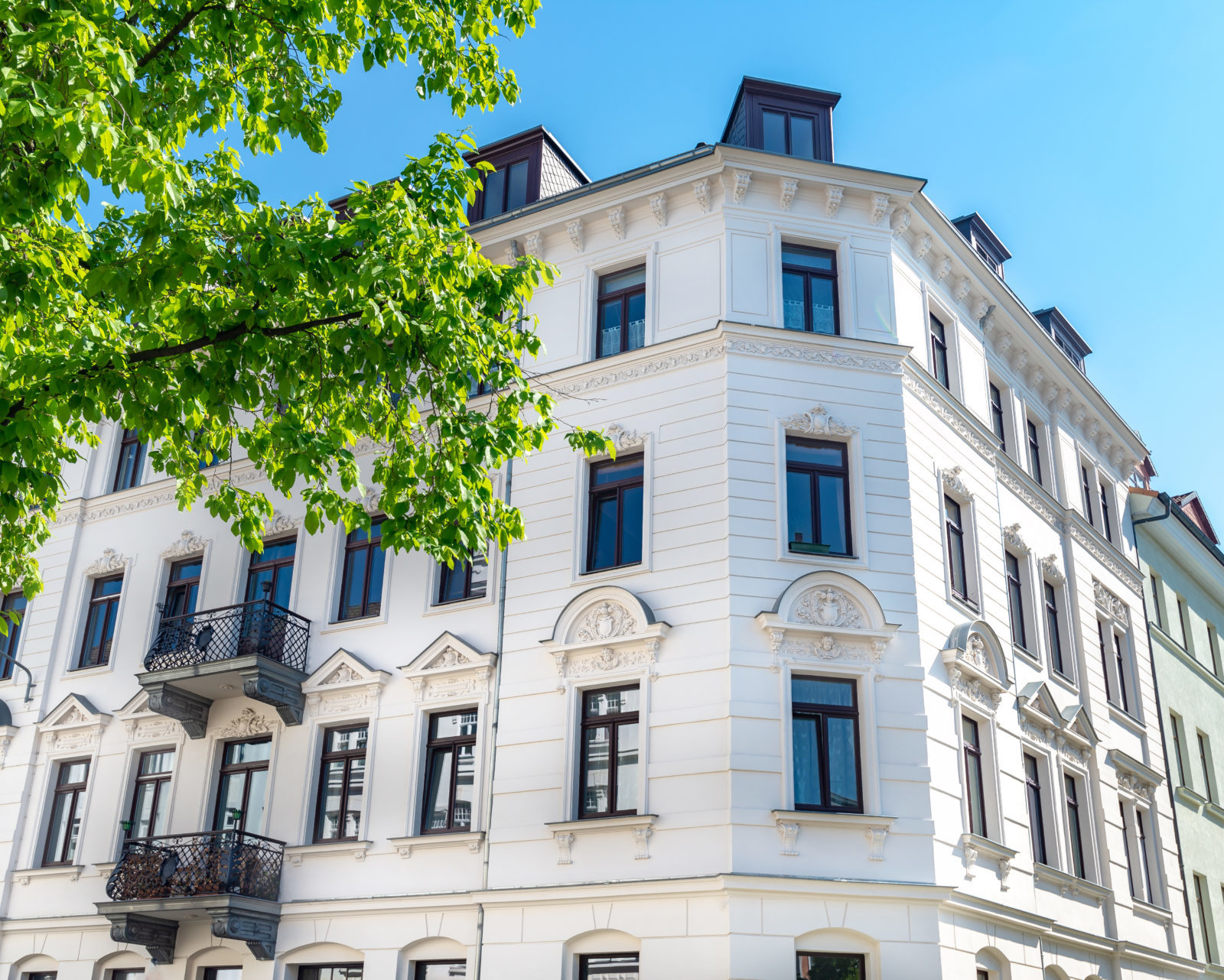 Leicht seitliche Frontansicht eines weißen Mehrfamilienhauses mit schwarzen Fensterrahmen und Stuckverzierung. Der Himmel ueber dem Haus ist hellblau und vor dem Haus ragt ein großer gruener Baum ins Bild.