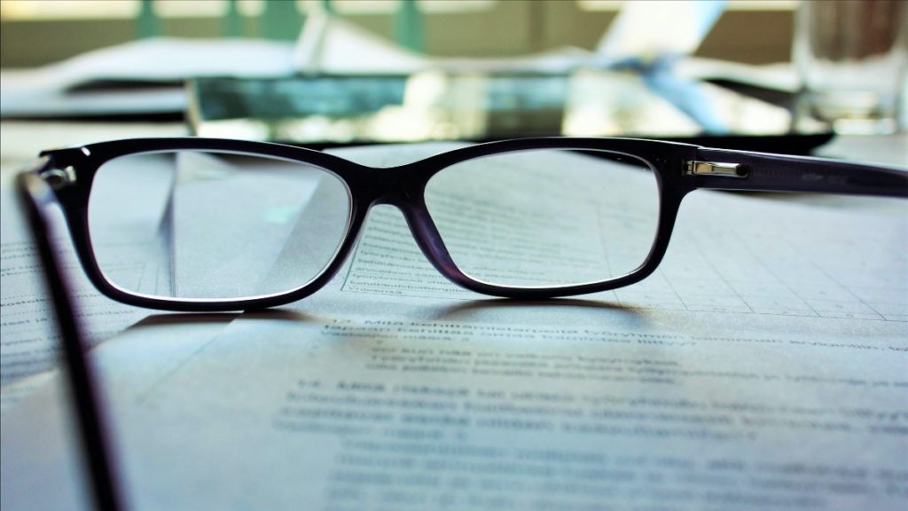 Ansicht einer Brille mit schwarzem, dickem Gestell, die auf mehreren Dokumenten zur Mietverwaltung liegt. Hinter der Brille und den Dokumenten sind nur unscharf zu erkennen ein Laptop und ein Glas.