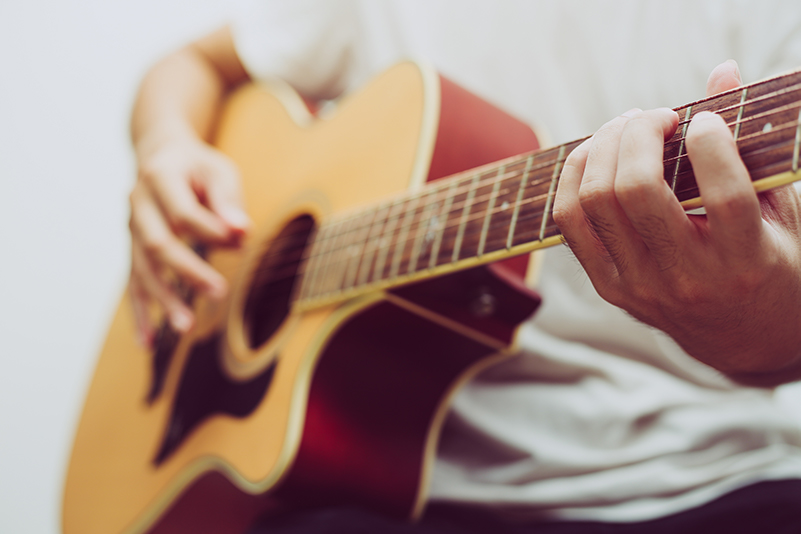 Mann mit weißem T-Shirt haelt eine braune Gitarre in den Haenden und hat seine Finger auf den Saiten.