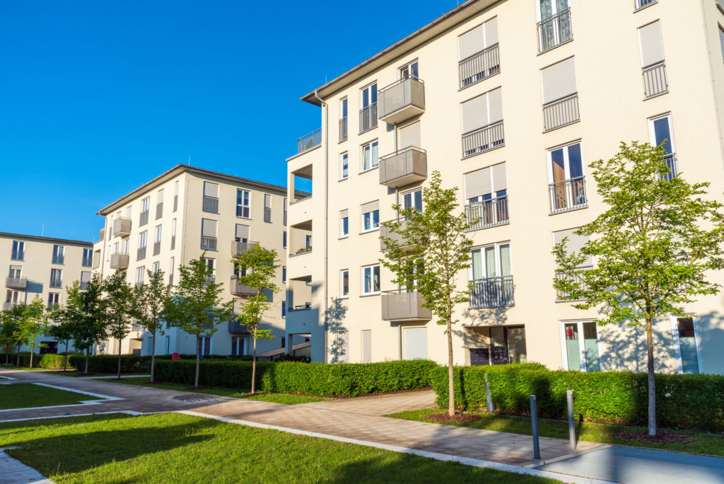 Leicht seitliche Ansicht mehrerer noch relativ neuer freistehender Mehrfamilienhaeuser mit einigen Balkonen und flachen Daechern. Vor den Haeusern befinden sich Gehwege, kleine Rasenflaechen und junge Baeume.