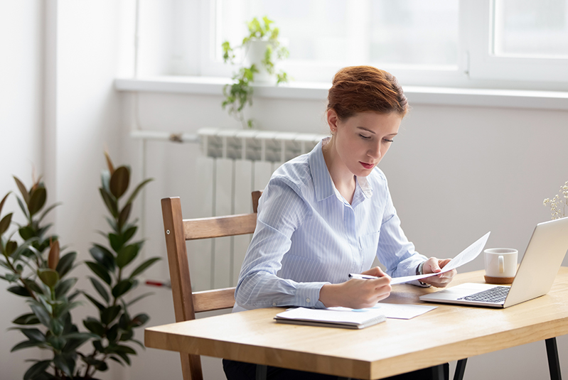 Junge Frau mit braun-roten Haaren und einer blau-weiß gestreiften Bluse sitzt an einem Schreibtisch. In der linken Hand haelt sie ein Dokument, in der rechten einen Stift. Neben ihr liegen weitere Dokumente. Vor ihr steht ein Laptop und im Hintergrund eine weiße Tasse.
