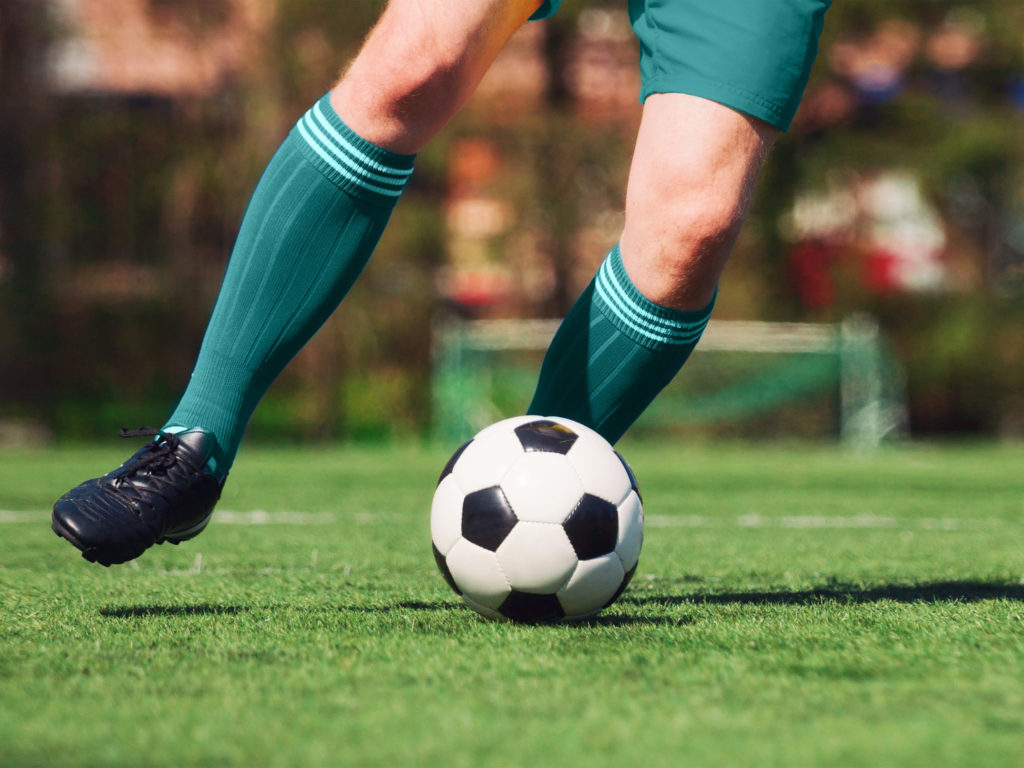 Beine eines Fußballers mit gruenen hochen Socken und schwarzen Stollenschuhen auf einem Rasen. Vor ihm liegt der Ball und er macht sich bereit zum Schuss. Im Hintergrund ist verschwommen ein Tor zu sehen.
