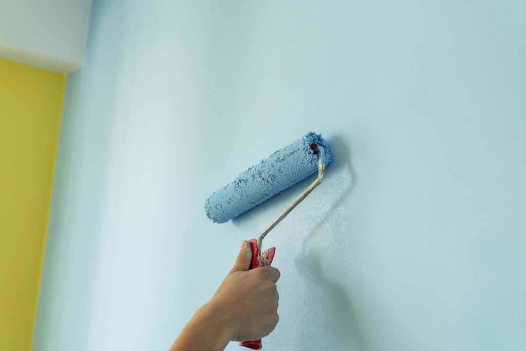 Hand mit Farbrolle in der Hand streicht eine Wand hellblau. Die Wand auf der linken Bildseite ist in einem kraeftigen gelb gestrichen.