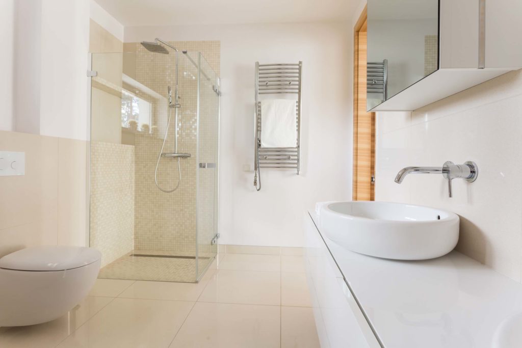 Ebenerdiges helles Badezimmer mit Glasdusche, weißem, runden Waschbecken auf einem langen Unterschrank und silbernem Wasserhahn und weißer Toilette gegenueber des Waschbeckens. Ueber dem Becken haengt ein Spiegelschrank. Neben der Dusche haengt eine Handtuchtrockner-Heizung an der Wand. Die Waende sind weiß und der Boden sowie die Seite von Dusche und WC beige.