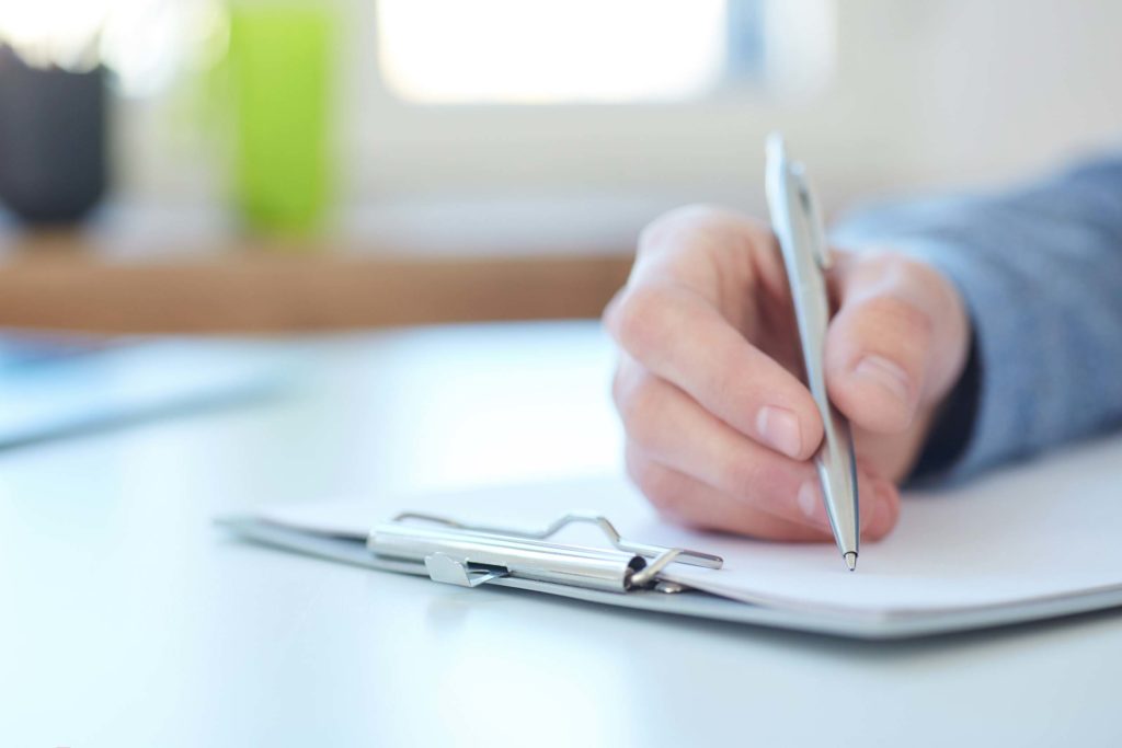 Hand einer Person mit blauem Hemd an einem weißen Tisch. Die Hand umfasst einen silbernen Stift, der sich auf einem weißen Klemmbrett mit mehreren Dokumenten befindet. Im Hintergrund ist eine Fensterbank mit Dekoelementen zu erahnen.