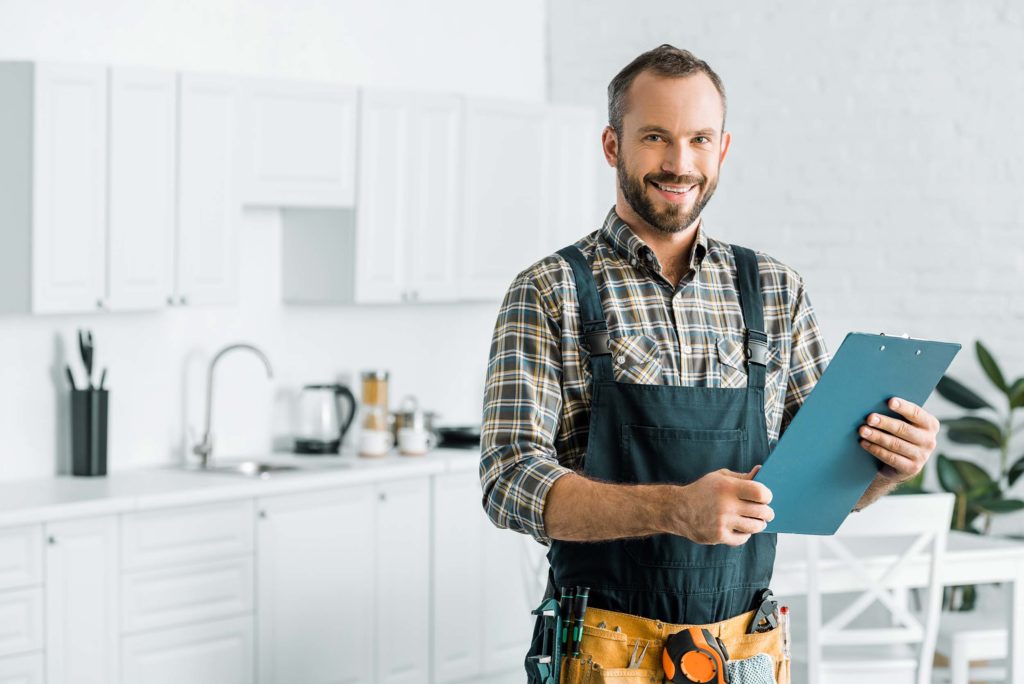 Mann mit kurzen Haaren und kariertem Hemd hat einen Overall an und haelt ein Klemmbrett in beiden Haenden. Er laechelt in die Kamera. In seinem Overall stecken verschiedene Werkzeuge wie Zangen und ein Maßband. Er steht in einer weißen Kueche mit weißen Schraenken, Tischen und Stuehlen. Kuechenzubehoer wir Kochloeffel, ein Wasserkocher und weiteres stehen im Hintergrund auf der Arbeitsplatte. Ein gruener Gummibaum steht im Hintergrund auf der rechten Seite hinter dem Esstisch.