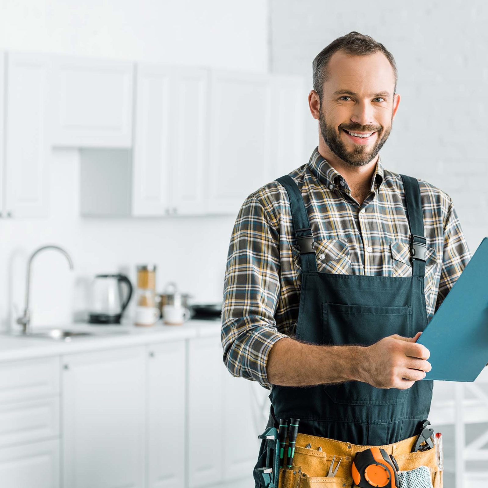 Mann mit kurzen Haaren und kariertem Hemd hat einen Overall an und haelt ein Klemmbrett in beiden Haenden. Er laechelt in die Kamera. In seinem Overall stecken verschiedene Werkzeuge wie Zangen und ein Maßband. Er steht in einer weißen Kueche mit weißen Schraenken, Tischen und Stuehlen. Kuechenzubehoer wir Kochloeffel, ein Wasserkocher und weiteres stehen im Hintergrund auf der Arbeitsplatte. Ein gruener Gummibaum steht im Hintergrund auf der rechten Seite hinter dem Esstisch.