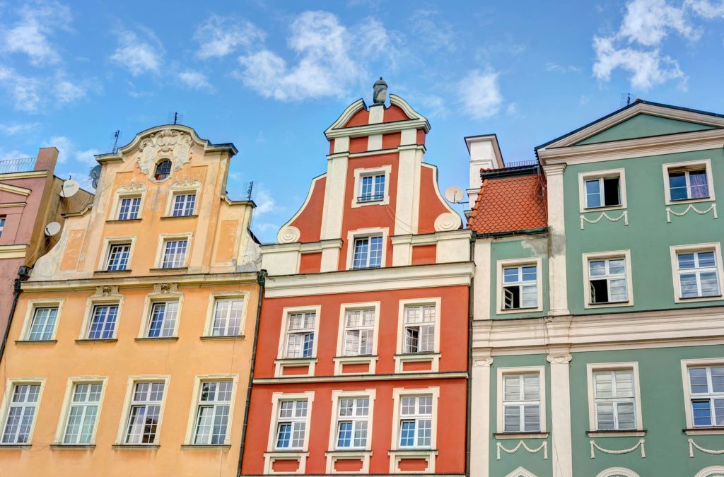 Denkmalschutz bei Frontansicht einer bunten Haeuserreihe in geld, rot und gruen mit unterschiedlichen Daechern, vielen Fenstern und Stuckverzierungen. Der Himmel ist blau mit wenigen weissen Wolken.