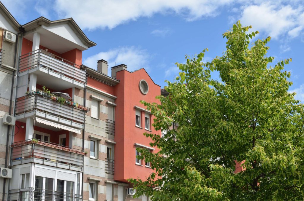 Leicht seitliche Ansicht einer roten Haeuserreihe mit Balkonen und vielen Fenstern. Vor den Haeusern waechst ein grosser gruener Baum und der Himmel ist blau mit wenigen weissen Wolken.