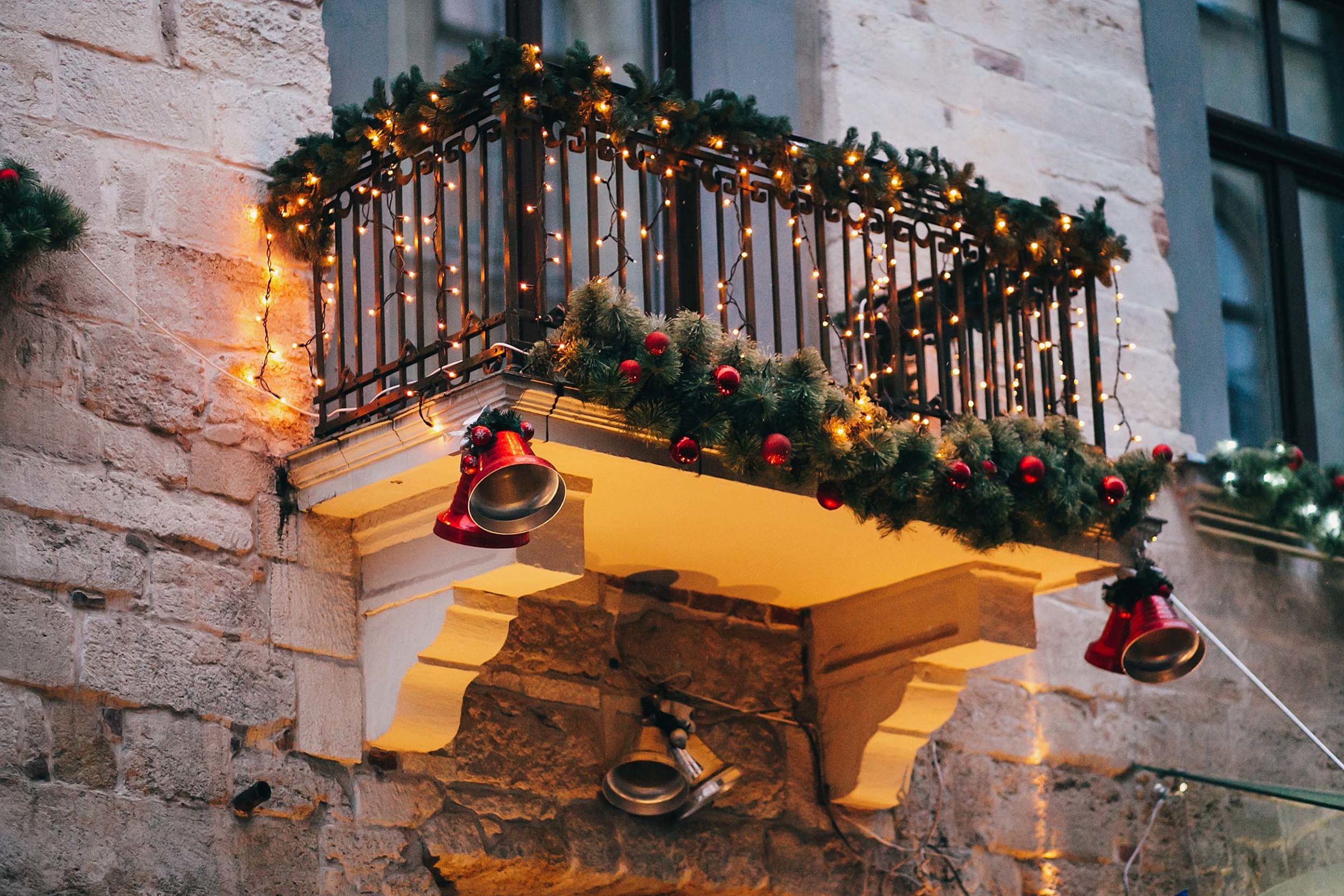 Weihnachtlich geschmueckter Balkon mit Tannengirlanden, Lichterketten und roten Christbaumkugeln. An den aeusseren Ecken das Balkons haengen jeweils zwei rote grosse Glocken. Mittig unter dem Balkon haengen zwei silberne Glocken. Die Abenddaemmerung setzt ein und die Lichterketten sind an.