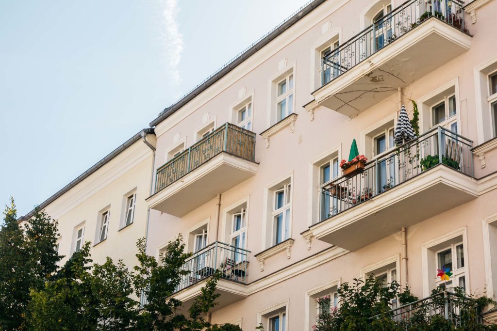Leicht seitliche Ansicht einer beigen Haeuserreihe mit mehreren Balkonen. Die Balkone sind mit Pflanzen, Schirmen und Kleinigkeiten dekoriert. Vor den Haeusern wachsen Baeume.
