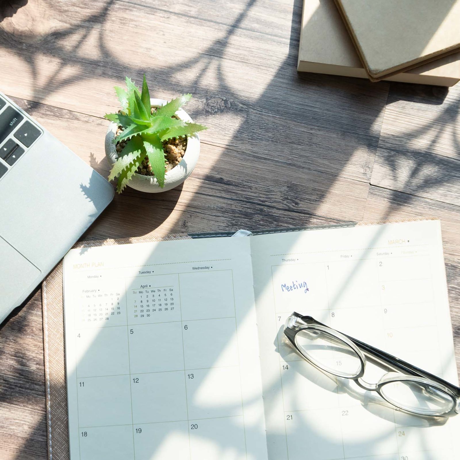 Blick auf einen Holzschreibtisch. Darauf liegen ein Laptop, ein geoeffneter Kalender, in dem in einem Kaestchen das Wort Meeting steht und ein Handy. Auf dem Kalender liegt eine Brille. Darueber steht eine kleine Pflanze. Im Hintergrund sind Teile von Buechern zu erkennen.