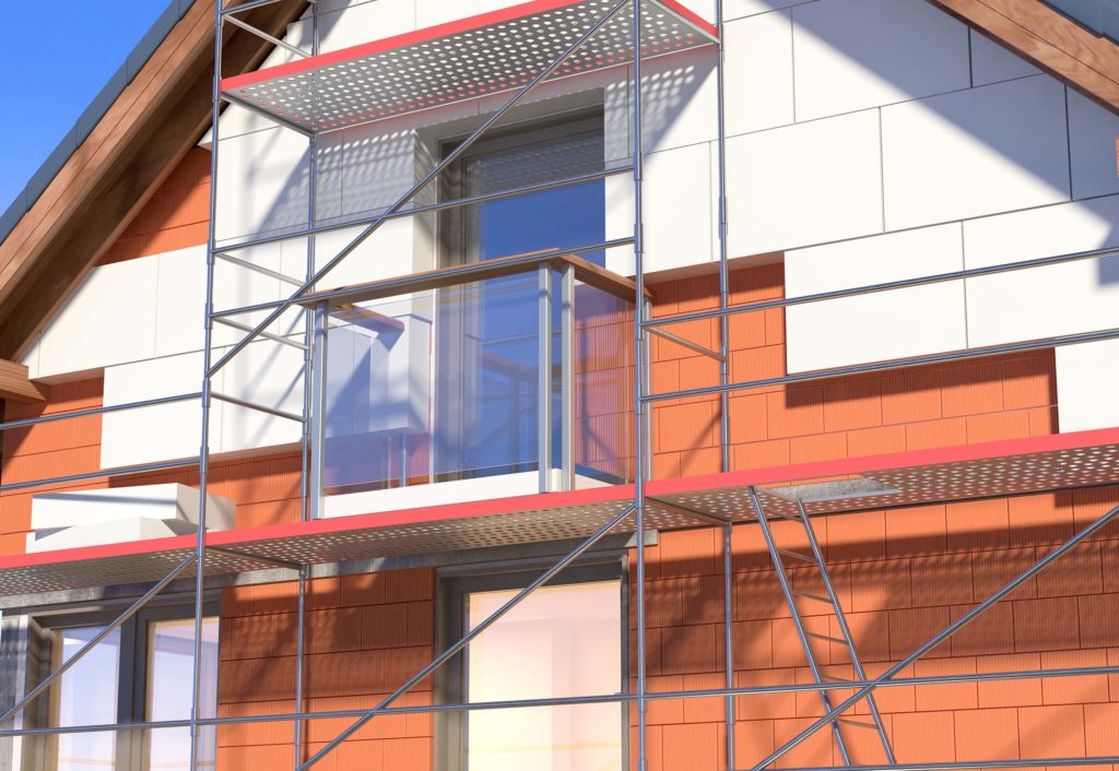Nahaufnahme eines Hauses im Bau. Der Rohbau ist rot und wird weiss verkleidet. Vor dem Haus ist ein Geruest aufgebaut. Innerhalb des Geruests wird ein Glasbalkon angebaut.