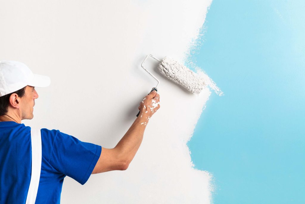 Handwerker mit blauem T-Shirt, weissem Overall und weissem Helm hat eine Farbrolle in der rechten Hand und streicht damit eine hellblaue Wand weiss. Die Wand ist auf der linken Seite bis circa zur Haelfte bereits gestrichen.