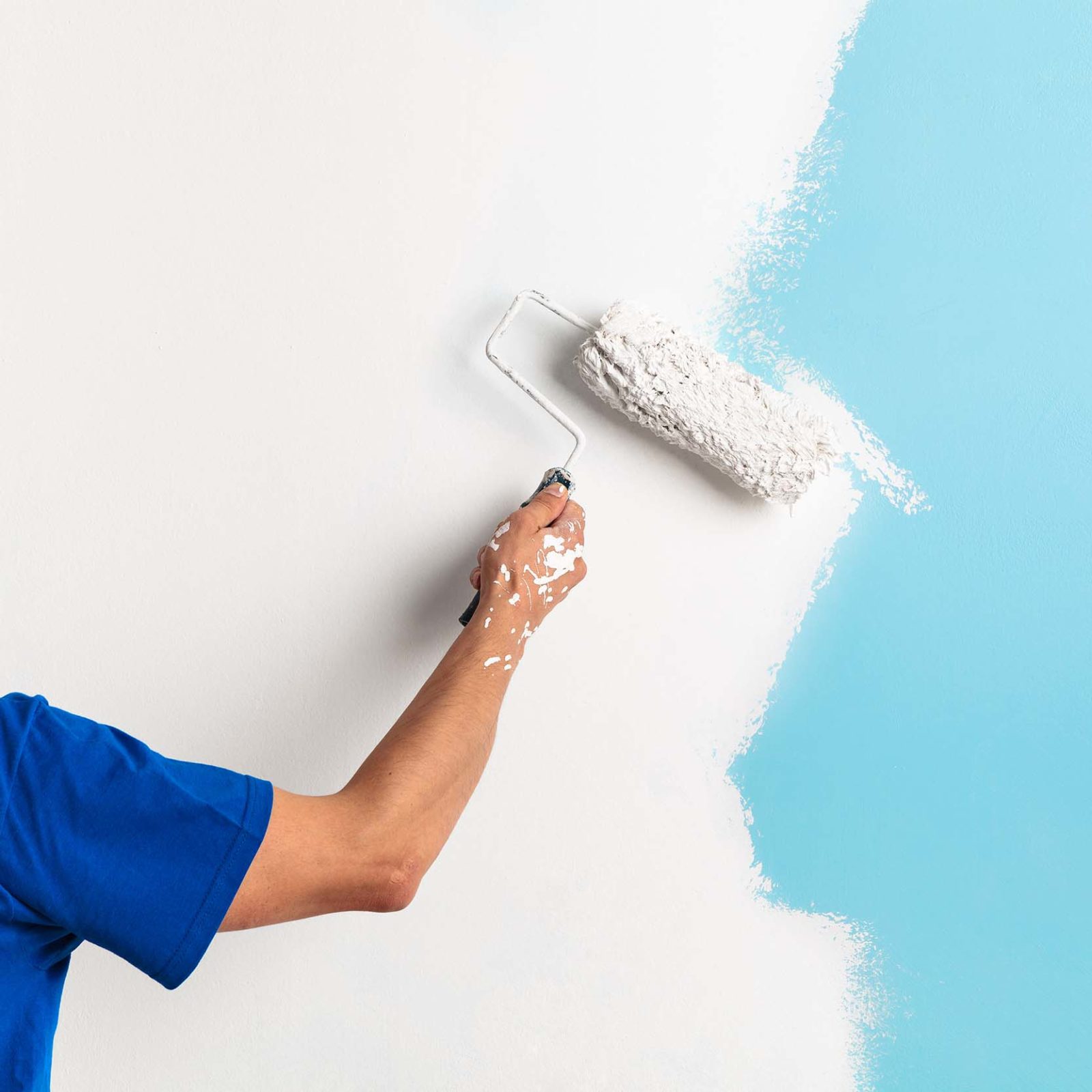 Handwerker mit blauem T-Shirt, weissem Overall und weissem Helm hat eine Farbrolle in der rechten Hand und streicht damit eine hellblaue Wand weiss. Die Wand ist auf der linken Seite bis circa zur Haelfte bereits gestrichen.