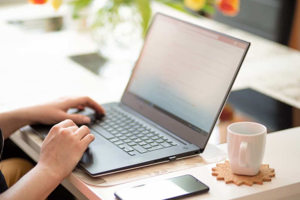 Haende einer Person, die an einem Schreibtisch sitzt. Die Haende liegen beide auf einem Laptop. Neben dem Laptop liegt ein Handy und eine weiße Tasse steht dort. Im Hintergrund ist eine Gruenpflanze zu erkennen.