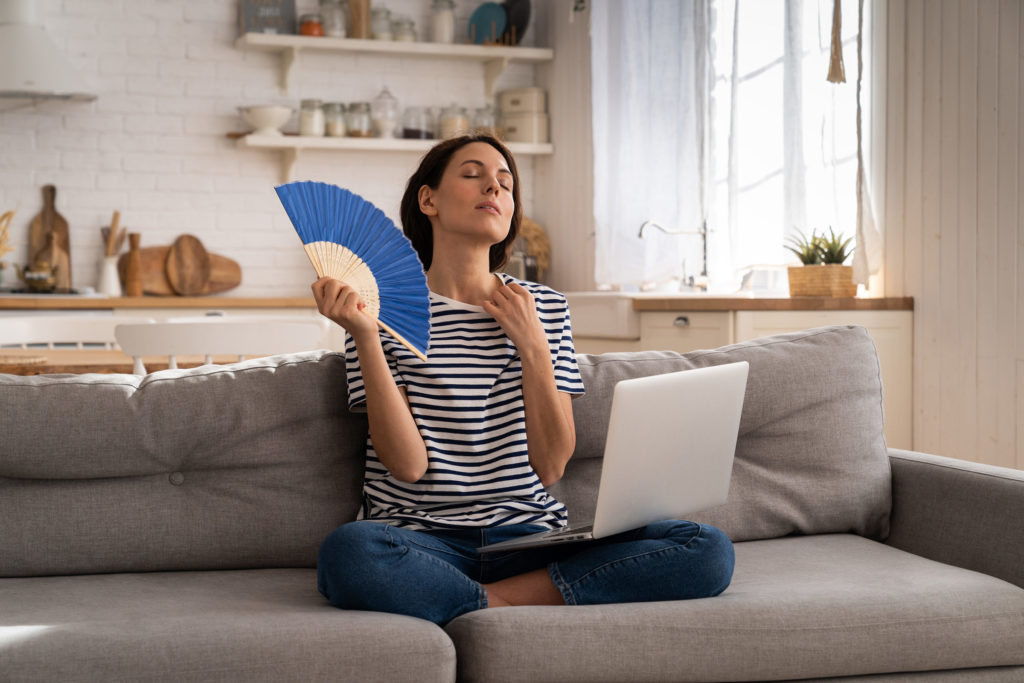Junge Frau mit braunen Haaren und gestreiftem T-Shirt sitzt mit einem Laptop auf dem Schoss auf einem grauen Sofa und faechert sich mit einem blauen Faecher Luft zu, waehrend sie mit der anderen Hand den Kragen ihres T-Shirts lockert, um sich abzukuehlen.