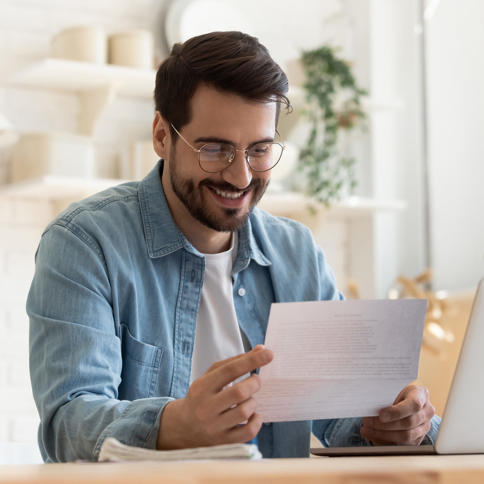 Nebenkostenabrechnung: Mann mit blauem Jeanshemd und weißem Shirt sitzt an einem Holztisch vor einem Laptop und haelt ein Dokument in den Haenden. Er laechelt dabei.