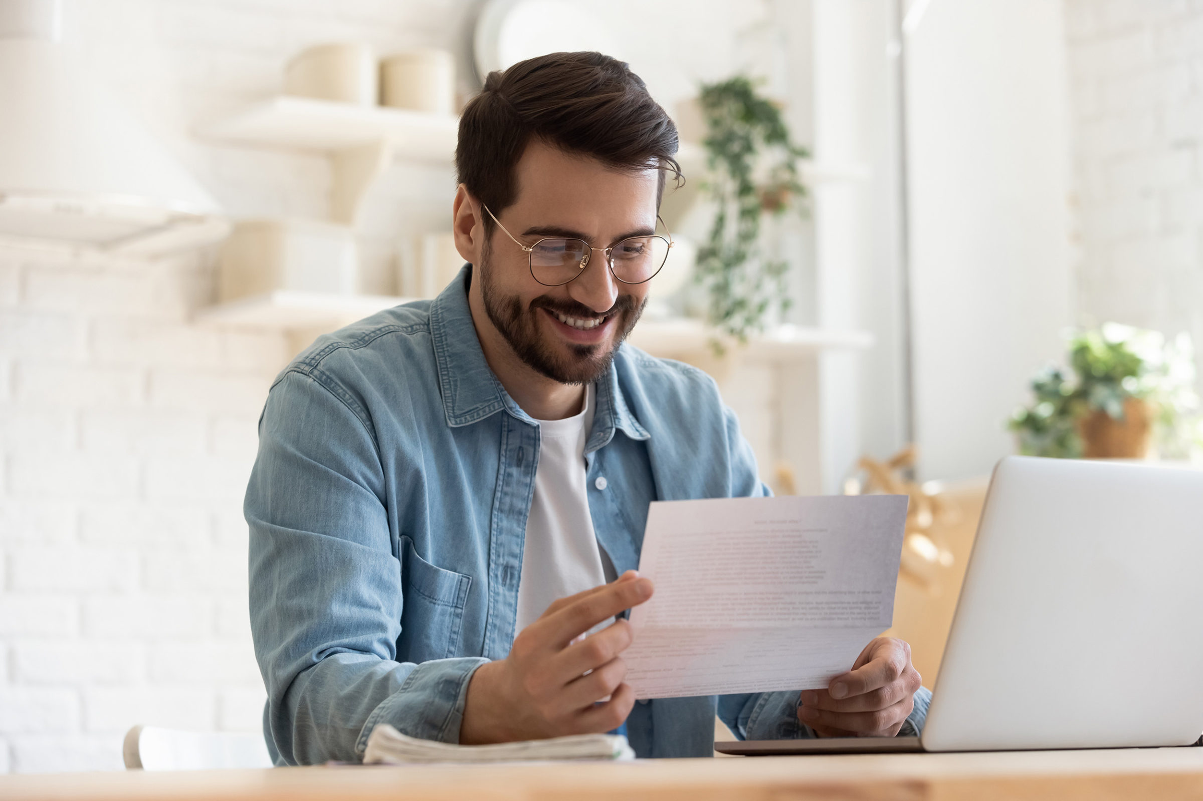 Nebenkostenabrechnung: Mann mit blauem Jeanshemd und weißem Shirt sitzt an einem Holztisch vor einem Laptop und haelt ein Dokument in den Haenden. Er laechelt dabei.