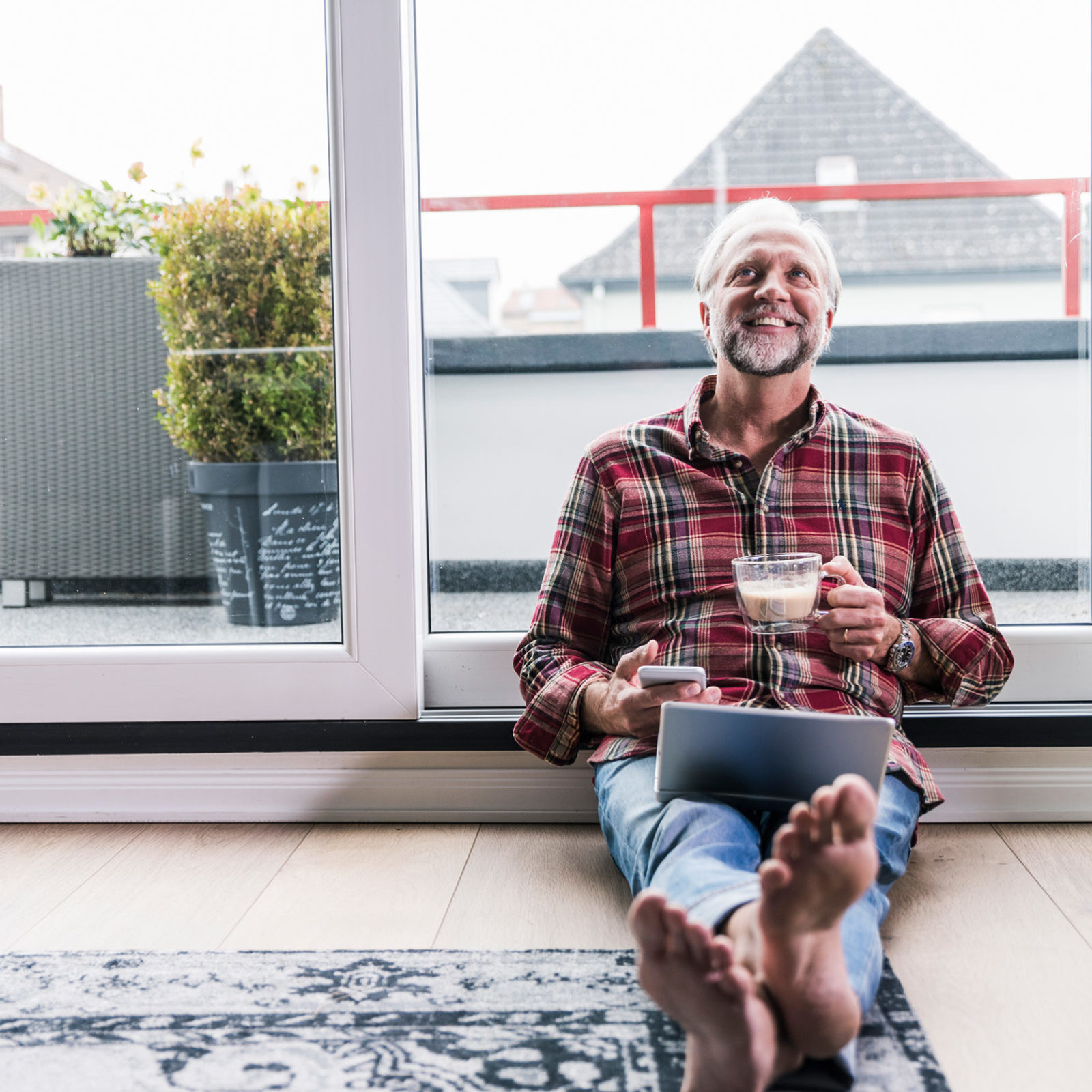 Mann mit kariertem Hemd sitzt auf dem Boden vor einem grossen Fenster. Er haelt eine Tasse in der Hand, auf seinem Schoss liegt ein Laptop und er streckt seine Fuesse Richtung Kamera. Auf dem Boden liegt ein großer Teppich. Auf dem Balkon hinter ihm stehen zwei Pflanzkuebel.