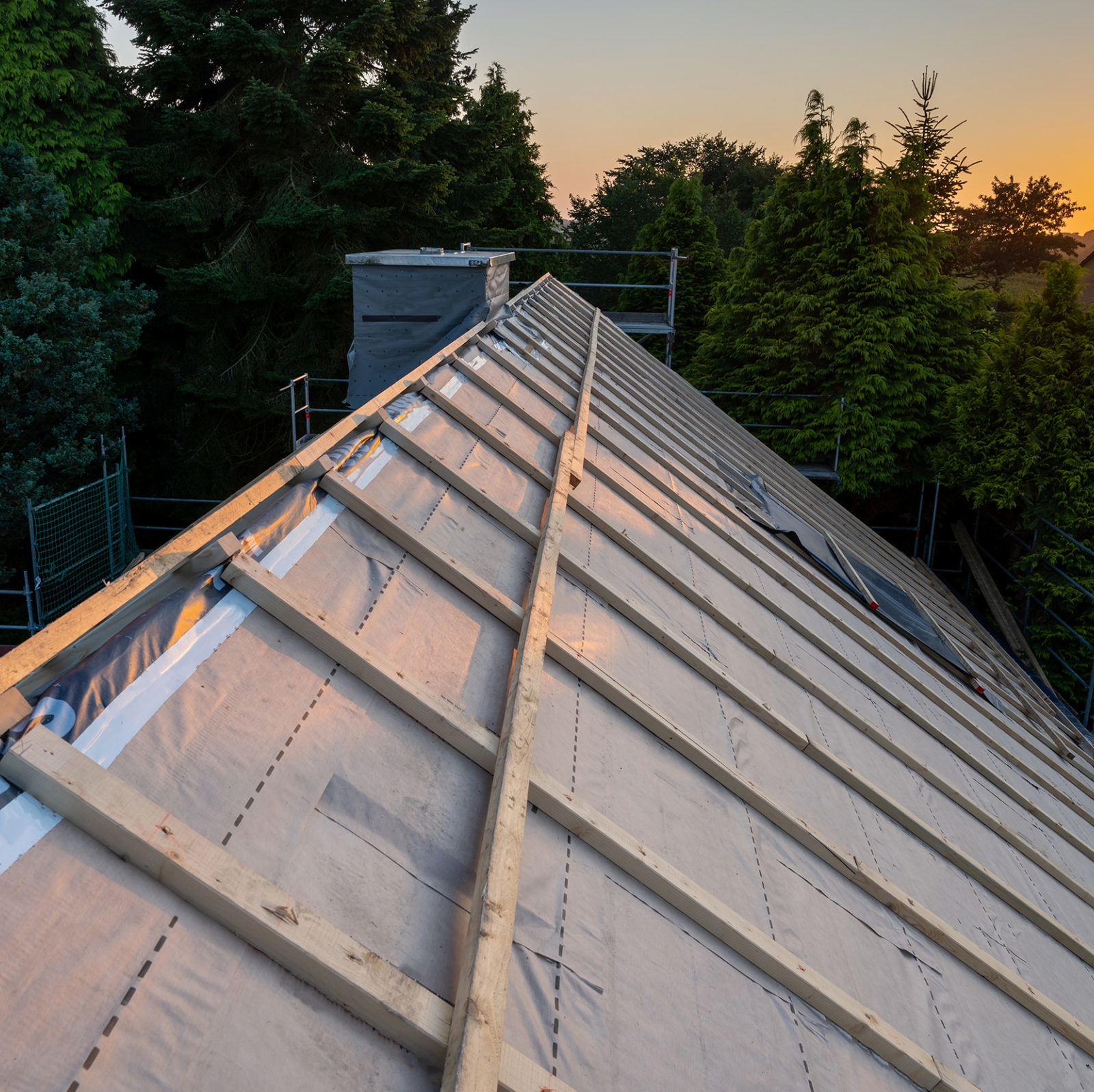 Ungedecktes Dach mit Holzbalken im Sonnenuntergang. Im Hintergrund sind Baeume.