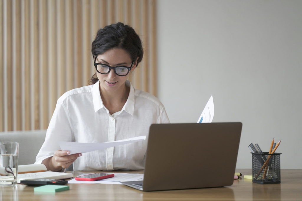 Junge Frau mit dunklen Haaren und weißer Bluse sitzt am Tisch und haelt Dokumente in den Haenden. Auf dem Tisch sind Laptop, Handy und Taschenrechner verteilt sowie weitere Dokumente.