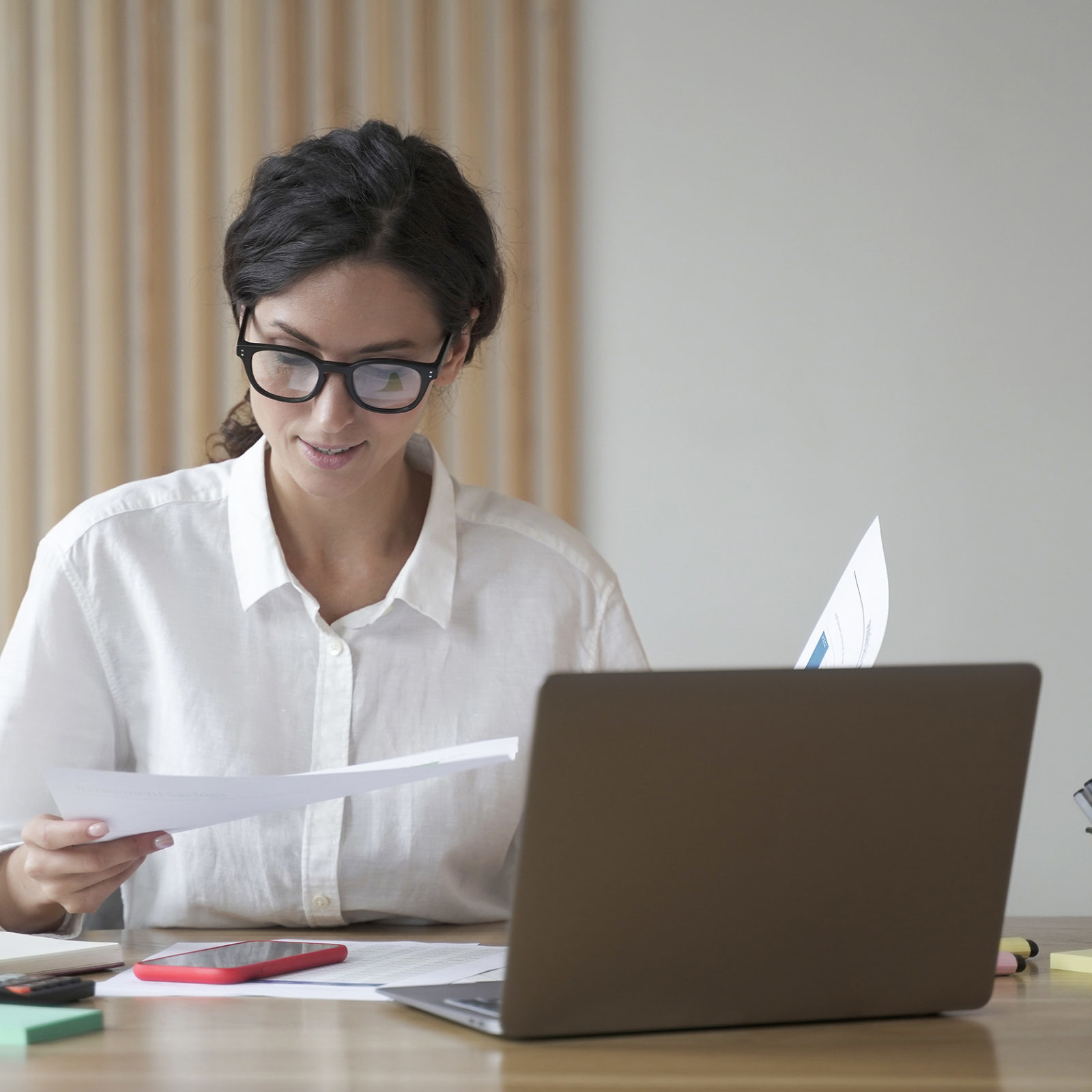 Junge Frau mit dunklen Haaren und weißer Bluse sitzt am Tisch und haelt Dokumente in den Haenden. Auf dem Tisch sind Laptop, Handy und Taschenrechner verteilt sowie weitere Dokumente.