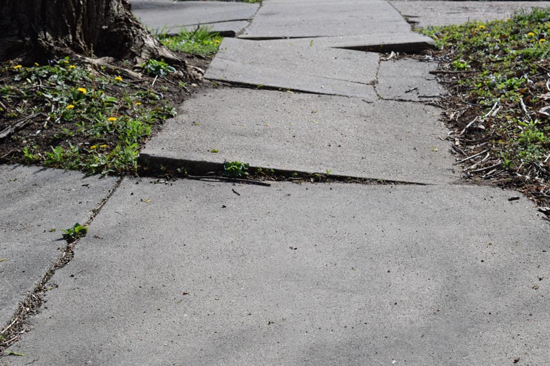 Verkehrssicherungspflicht: Gehweg aus quadratischen Platten, die Hoehenunterschiede haben. An den Seiten waechst Gras.