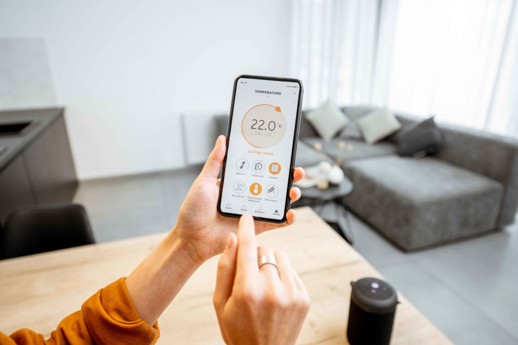 Person mit orangefarbenem Oberteil haelt ein Handy mit einer Hand hoch und tippt mit der anderen Hand darauf. Auf dem Bildschirm ist die Steuerung einer Heizung zu sehen, die auf zweiundzwanzig Grad eingestellt ist. Im Hintergrund sind ein Holztisch, auf dem ein mobiler Lautsprecher steht, ein graues Sofa, eine dunkelgraue Kueche und ein kleiner Wohnzimmertisch zu sehen.