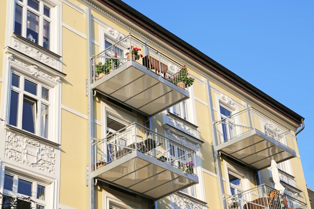 Leicht seitliche Ansicht eines gelben Mehrfamilienhauses mit Stuckverzierung und nachtraeglich vor der Fassade angebrachten silbernen Balkonen. Die Balkone sind mit Pflanzen und Schirmen dekoriert. Der Himmel ist blau.