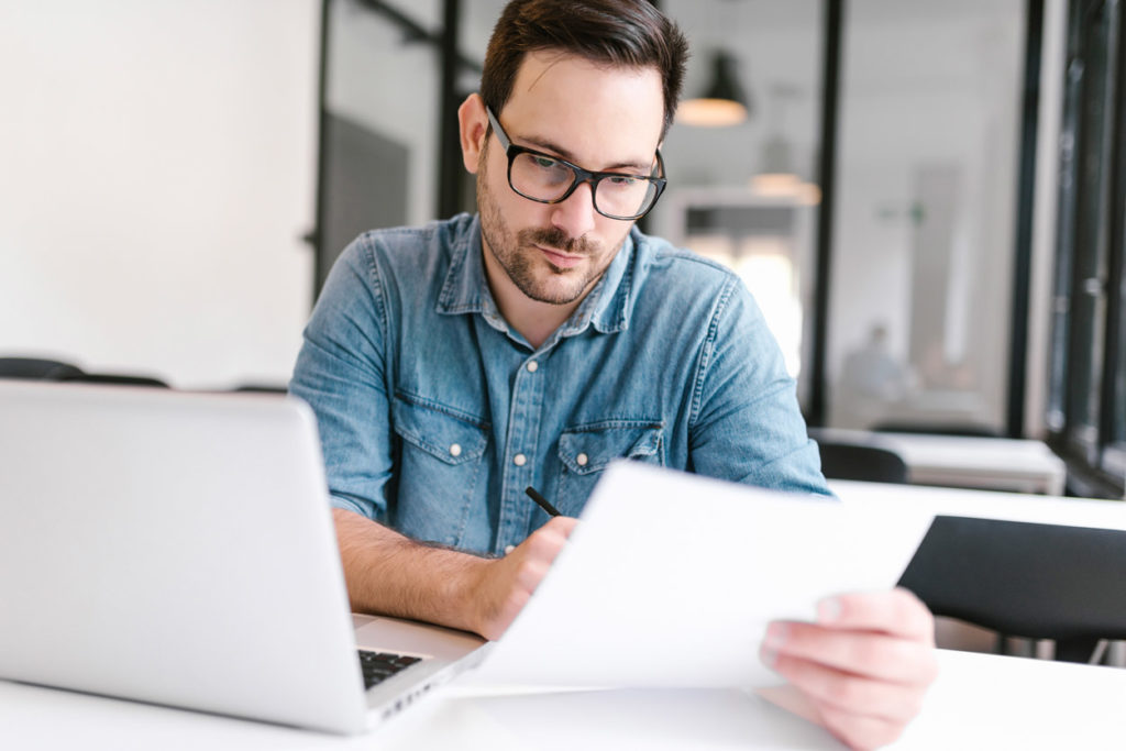 Mann mit kurzen dunklen Haaren, Brille und blauem Jeanshemd sitzt an einem Tisch und haelt mit einer Hand ein Dokument, welches er liest. In der anderen Hand hat er einen Stift. Vor ihm steht ein Laptop. Im Hintergrund spiegelt sich der Mann. An der Decke haengt eine schwarze Lampe.