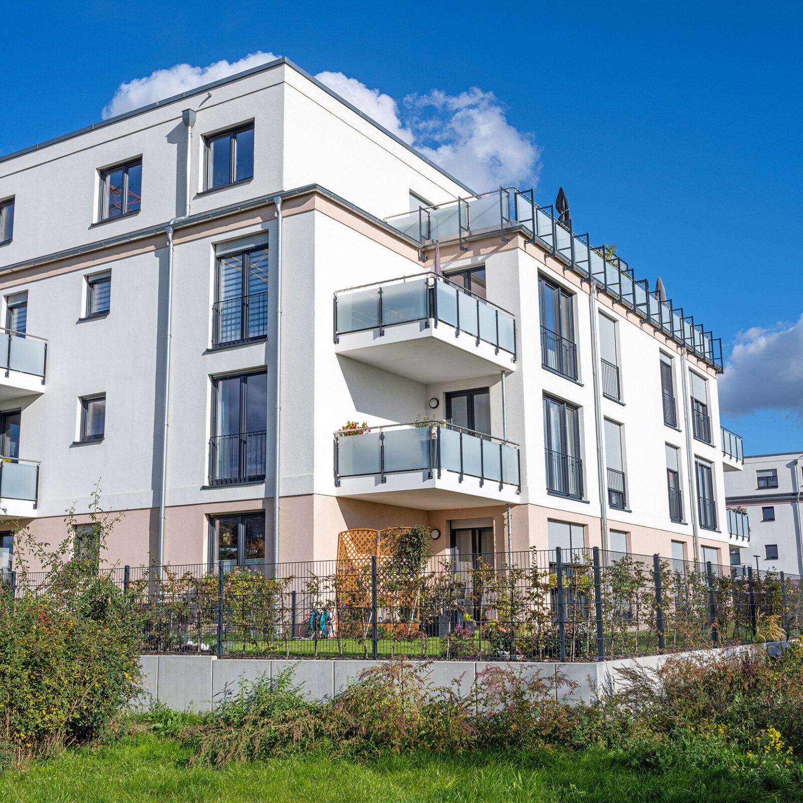 Leicht seitliche Ansicht auf mehrere freistehende Mehrfamilienhaeuser mit dunklen Fensterrahmen und vorgesetzten Balkonen. Die Balkone sind zum Teil bepflanzt. Die Haeuser haben flache Daecher und sind umzaeunt. Vor den Zaeunen befindet sich eine Rasenflaeche mit wild wachsenden Pflanzen. Der Himmel im Hintergrund ist blau mit vereinzelten Wolken.