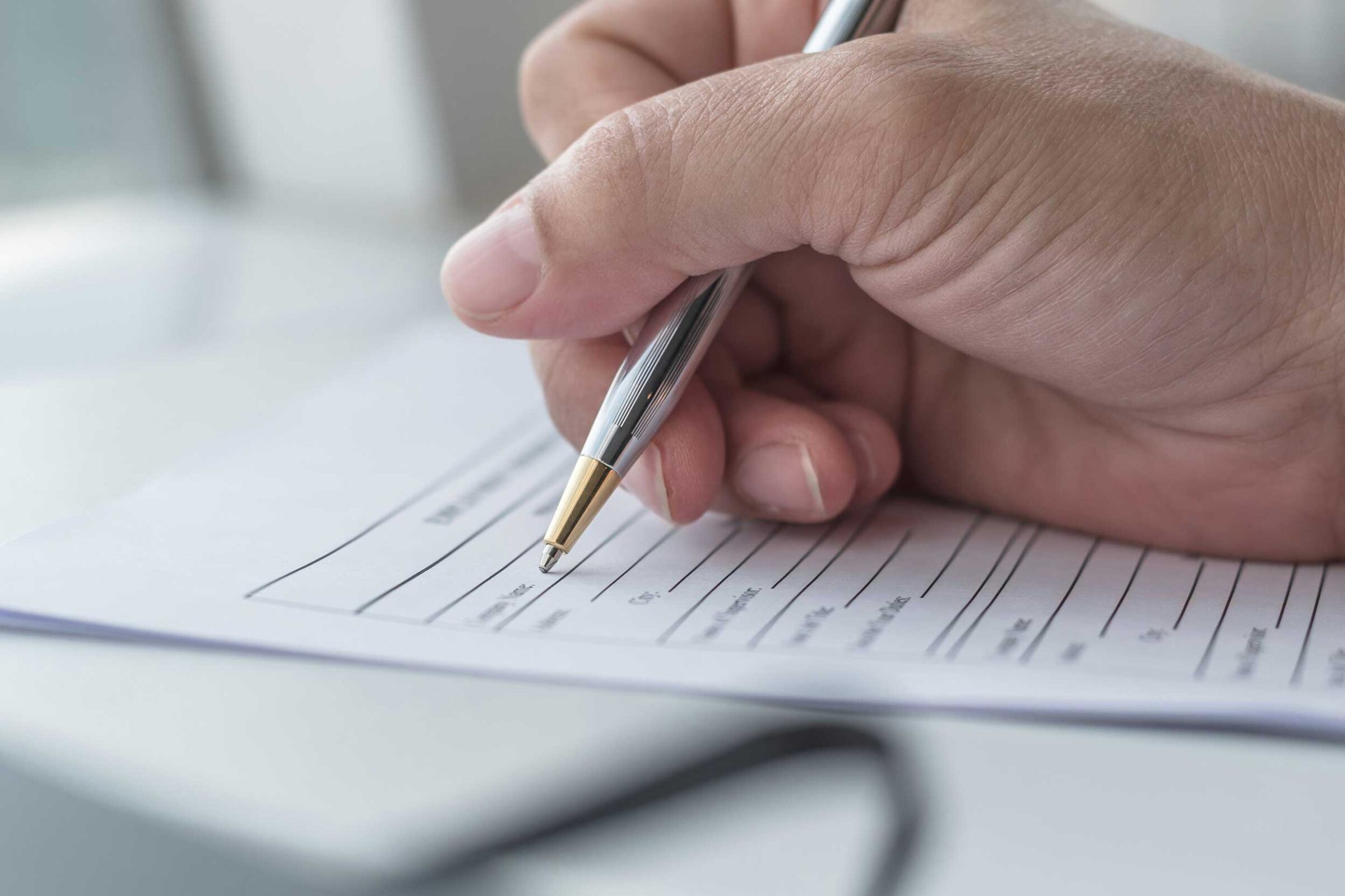 Hand, die einen Stift umfasst, setzt zum Schreiben auf einem Dokument an, das auf einem weißem Tisch liegt.