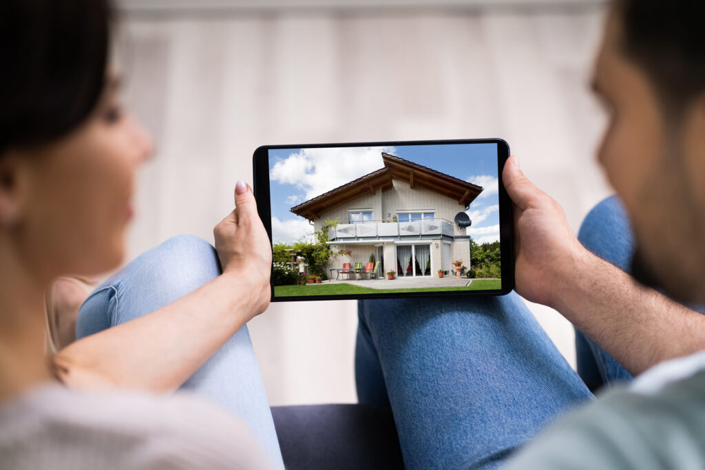 Online Wohnungsbesichtigung: Ein Paar sitzt auf einer Couch. Die Frau links und der Mann rechts. Zusammen halten sie ein Tablet in den Haenden, auf dem die Aussenansicht zum Garten hin eines Hauses zu sehen ist. Der Fokus liegt auf dem Tablet, die Personen sind nur zu einem geringen Teil zu sehen und verschwommen. Der Boden im Hintergrund ist hell.