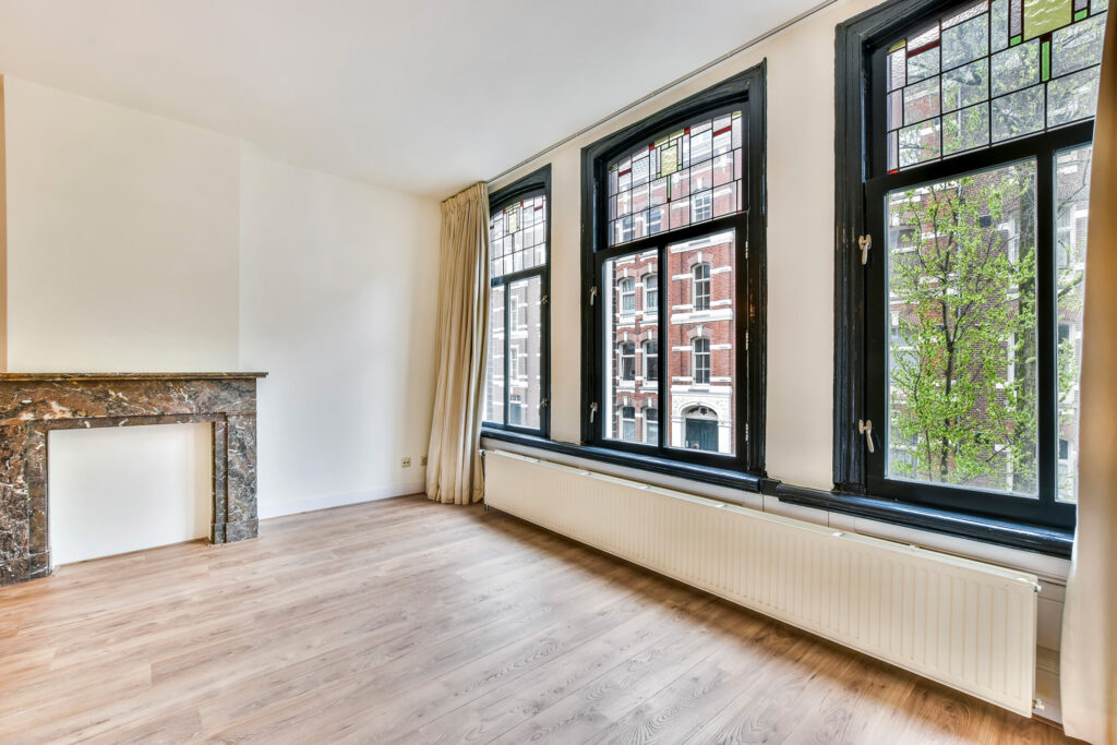 Wohnungsleerstand: Schraeger Blick in ein leeres Zimmer einer Wohnung. Auf der rechten Seite ist eine grosse Fensterfront aus drei Fensterlementen und jeweiligem Oberlicht. Die Fenster haben dunkle Rahmen und die niedrige Fensterbank hat den selben Farbton. Darunter befindet sich ein langer, schmaler Heizkoerper, der fast ueber die gesamte Zimmerlaenge geht. An den Seiten sind beige Vorhaenge hoch angebracht, die ueber die gesamte Zimmerhoehe gehen. Die Waende sind weiss und der Boden aus hellem Holz mit Maserung. Auf der linken Bildseite ist ein Kaminsims, aber ohne Kamin.