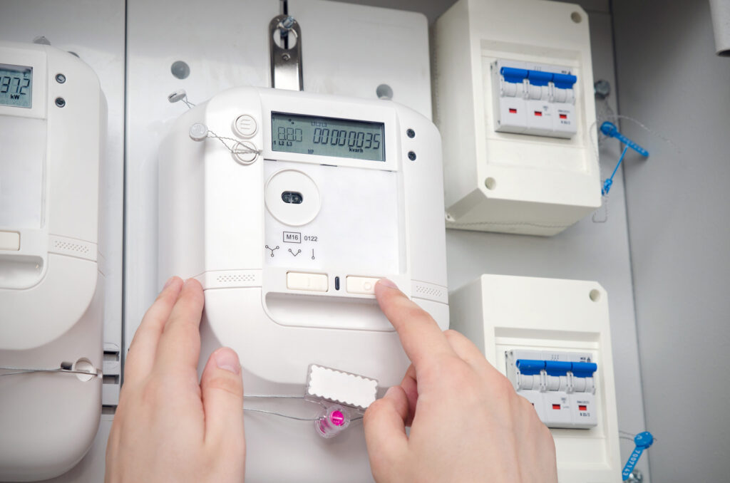 Smart Meter Gateway: Leicht seitlicher Blick auf digitale Zaehler. Ein Zaehler ist im Vordergrund. Die Zaehler sind weiss. Die Haende einer Person sind im unteren Bildabschnitt zu sehen. Die linke Hand beruehrt den Zaehler mit drei Fingern. Der Zeigefinger der rechten Hand beruehrt einen der Knoepfe auf dem Zaehler. Rechts neben dem Zaehler sind zwei kleine Sicherungskaesten mit jeweils drei Sicherungen angebracht.