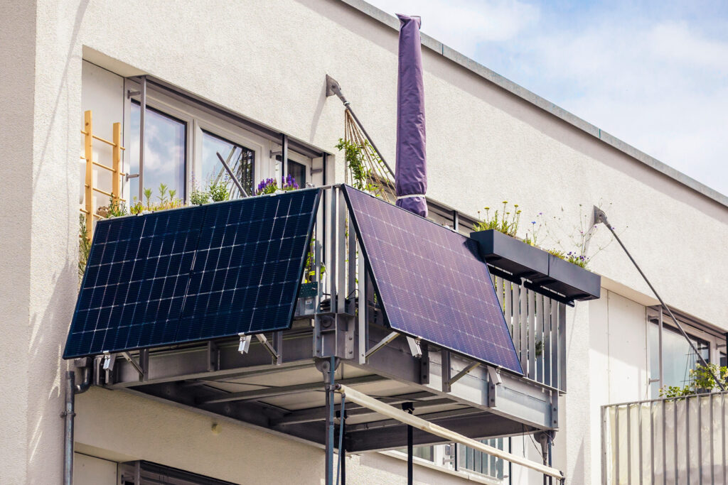 Balkonkraftwerk: Seitlicher Blick, leicht von unten auf einen Balkon an einem weissen Haus. Der Balkon hat ein silbernes Gelaender an dem frontal und an der einen Seite jeweils ein dunkelblaues Solarpanel angebracht ist. Auf dem Balkon steht ein geschlossener Sonnenschirm mit einer dunkelblauen Huelle. Am Gelaender des Balkons sind rundum Blumen gepflanzt. Im Hintergrund sind Fenstertueren und die Hauswand zu sehen.