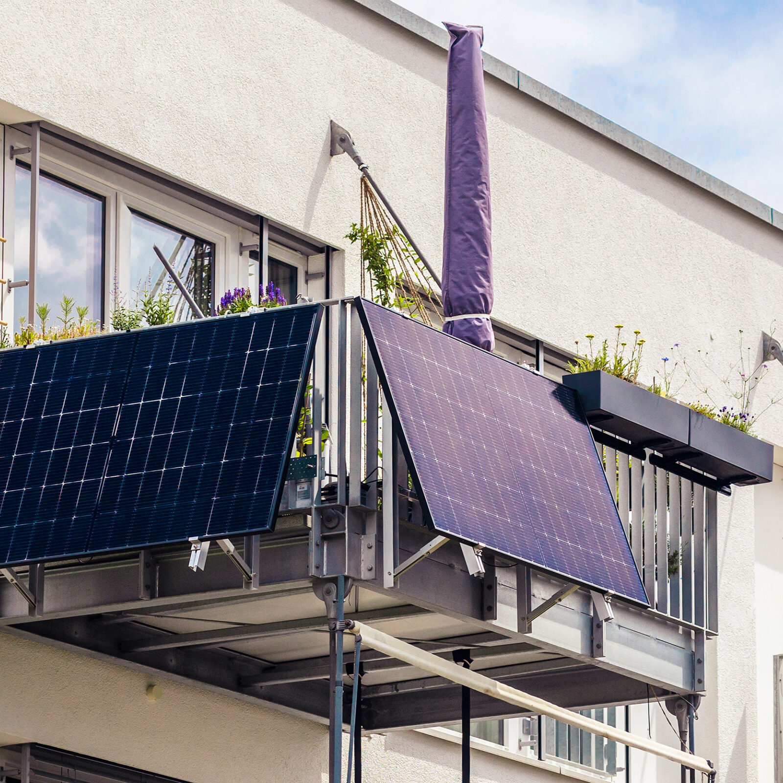 Balkonkraftwerk: Seitlicher Blick, leicht von unten auf einen Balkon an einem weissen Haus. Der Balkon hat ein silbernes Gelaender an dem frontal und an der einen Seite jeweils ein dunkelblaues Solarpanel angebracht ist. Auf dem Balkon steht ein geschlossener Sonnenschirm mit einer dunkelblauen Huelle. Am Gelaender des Balkons sind rundum Blumen gepflanzt. Im Hintergrund sind Fenstertueren und die Hauswand zu sehen.