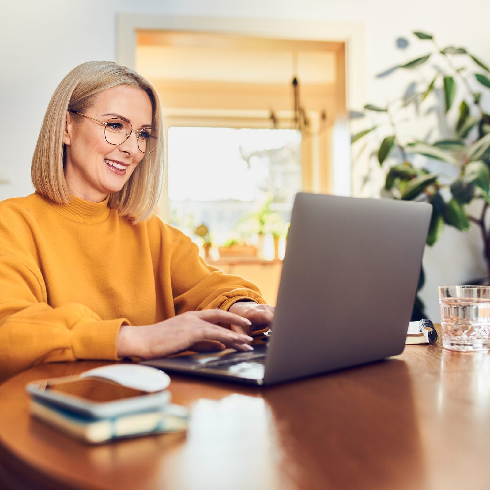 Aenderungen 2025: Frau mit blonden, schulterlangen Haaren, Brille und gelbem Pullover sitzt an einem dunklen Holztisch vor einem Laptop. Ihre Haende sind auf dem Laptop abgestützt und die Finger ruhen auf den Tasten. Sie laechelt beim Blick auf den Bildschirm. Neben dem Laptop liegen mehrere Notizbuecher. Im Hintergrund steht eine groesse Gruenplfanze.