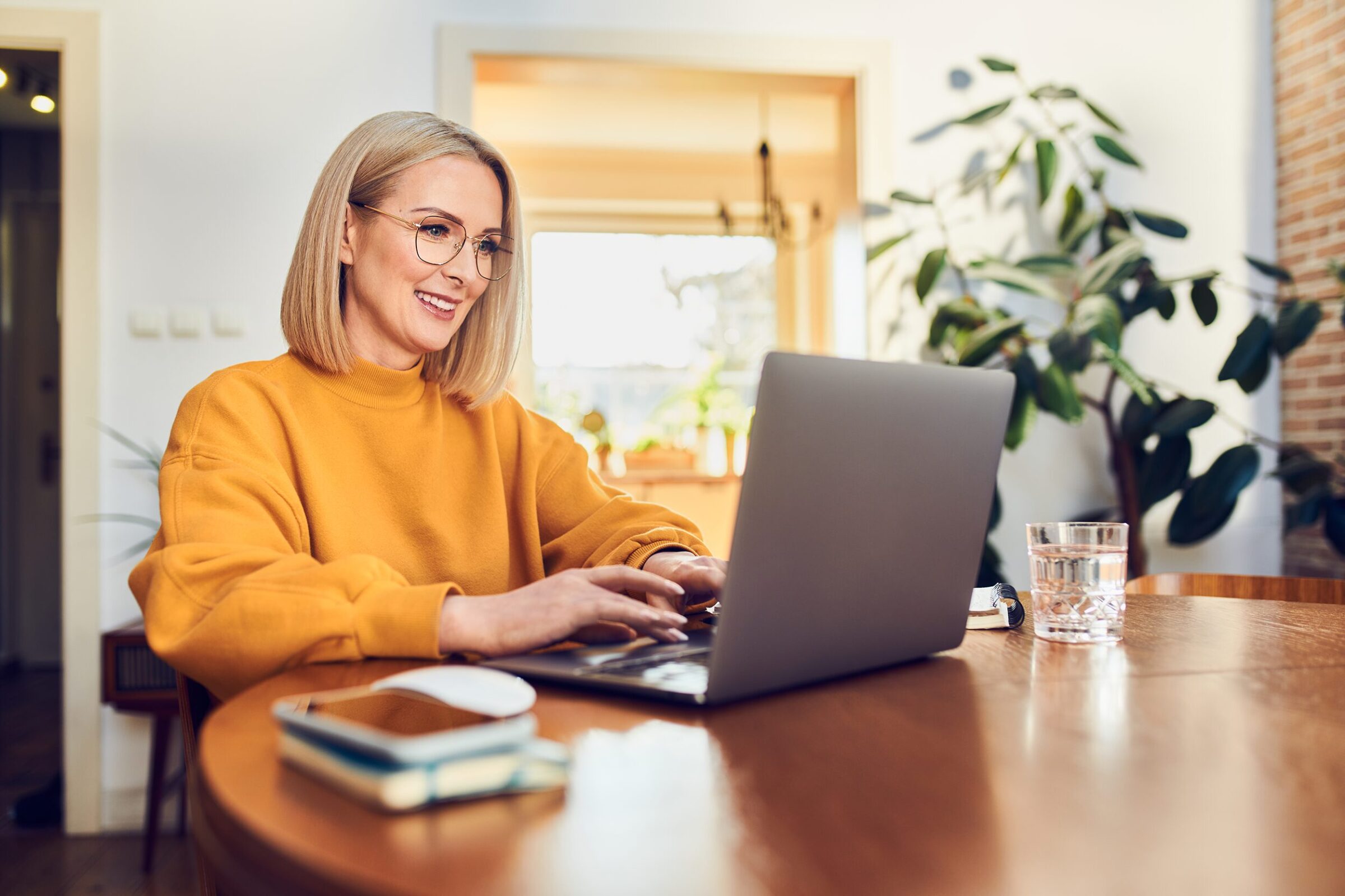 Aenderungen 2025: Frau mit blonden, schulterlangen Haaren, Brille und gelbem Pullover sitzt an einem dunklen Holztisch vor einem Laptop. Ihre Haende sind auf dem Laptop abgestützt und die Finger ruhen auf den Tasten. Sie laechelt beim Blick auf den Bildschirm. Neben dem Laptop liegen mehrere Notizbuecher. Im Hintergrund steht eine groesse Gruenplfanze.