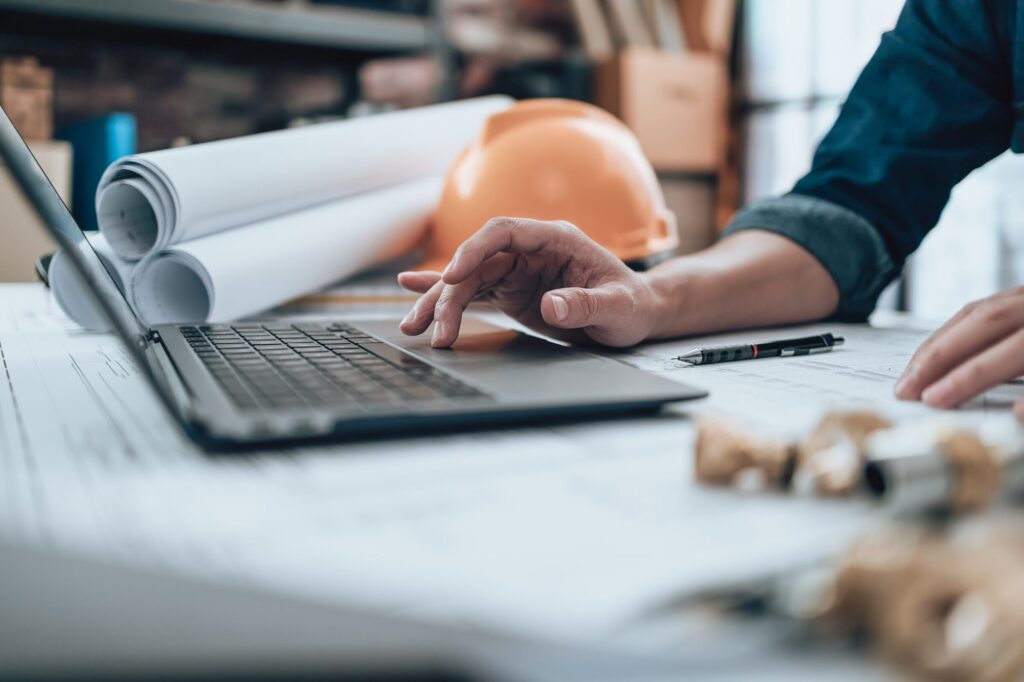 Verringerung der Betriebskosten durch Modernisierung: Nahaufnahme eines Tisches. Darauf liegen zahlreiche Dokumente und ein aufgeklappter Laptop steht auf diesen Dokumenten. Vom rechten Bildrand ist ein Teil einer Person mit dunkelblauem Jeanshemd zu sehen. Die rechte Hand ist vor dem Laptop abgestuetzt und ein Finger ruht auf dem Trackpad.Im Hintergrund sind drei grosse Papierrollen zu sehen, daneben ein orangener Sicherheitshelm.