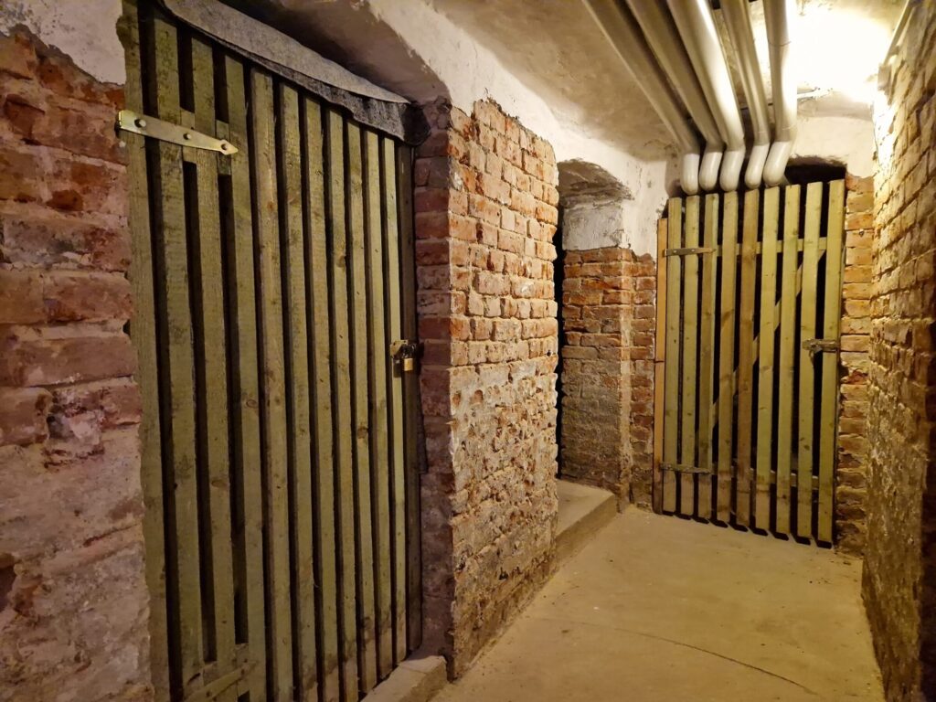 Feuchtigkeit im Keller von Altbauwohnung: Leicht seitlicher Blick auf einen Kellerraum mit Tueren aus Holzbalken. Dazwischen befinden sich Backsteinmauern, der Boden besteht aus Beton.