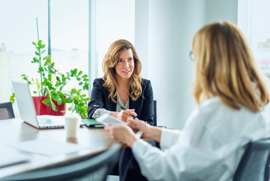 Mietverwaltung: Zwei Frauen mit dunkelblonden mittellangen Haaren sitzen an einem Tisch in einem hellen Raum. Sie sprechen miteinander. Auf dem Tisch stehen ein Laptop, ein kleiner Ordner, einzelne Dokumente und ein To-Go Kaffeebecher. Im Hintergrund steht eine Gruenpflanze.